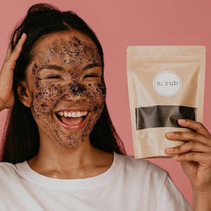 woman with coffee scrub on face holding packet of scrub